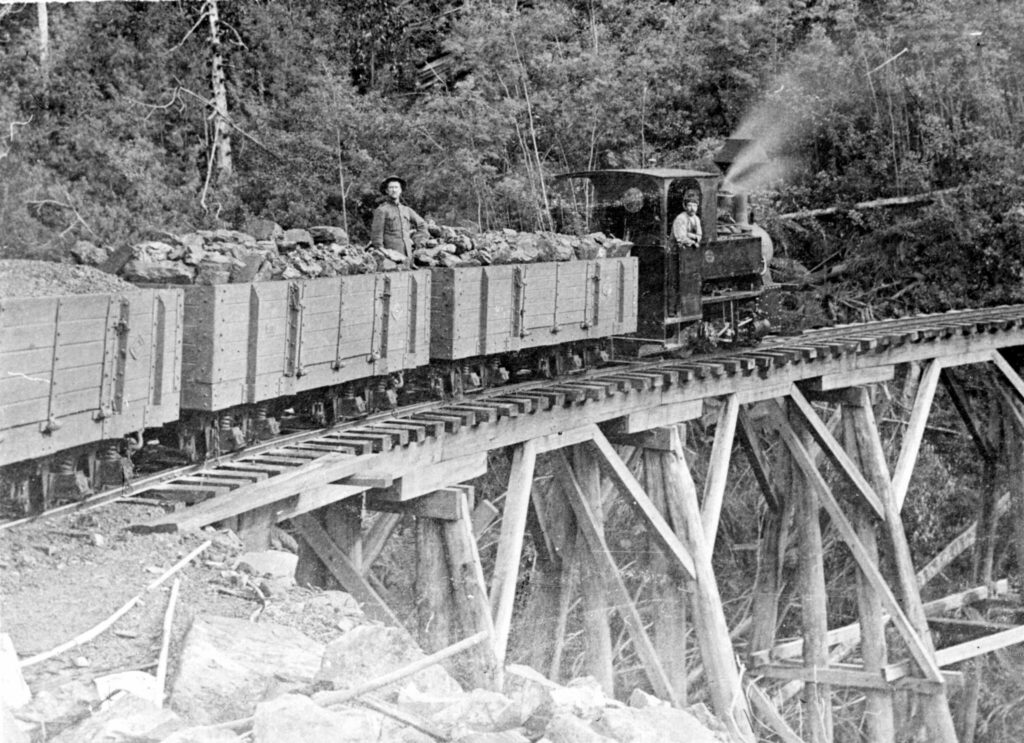 Loaded coal train heading down hill on the Sandfly Colliery Tramway tailed by the 2-4-0T Krauss locomotive. This line ran from coal mines at Kaoota, in the hills around Sandfly, to Margate, south of Hobart on North West Bay, for shipping. Ted Lidster collection via Flickr user Traniac - https://www.flickr.com/photos/29903115@N06/11668439343/in/photostream/
